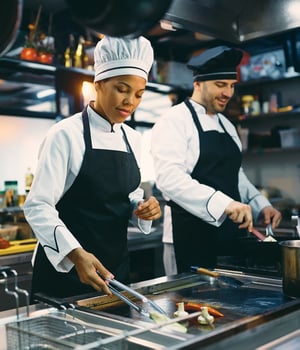 Two chefs happily preparing meals in a restaurant kitchen. On the left, a female chef wearing a white shirt, black apron, and white hat; on the right, a male chef in matching attire. The seamless restaurant management results from tamigo's highly effective restaurant workforce management software.