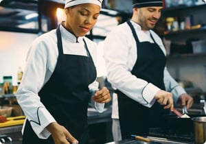 Two chefs happily preparing meals in a restaurant kitchen. On the left, a female chef wearing a white shirt, black apron, and white hat; on the right, a male chef in matching attire. The seamless restaurant management results from tamigo's highly effective restaurant workforce management software.