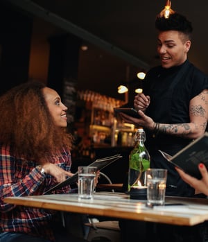 Cheerful male waiter with arm tattoos, wearing a black t-shirt, joyfully taking the meal order of a delighted woman dressed in a red and blue flannel shirt. The restaurant's efficient operations owe credit to advanced restaurant management software.