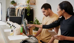 Assistentes de vendas eficientes, homens e mulheres, a trabalharem em conjunto, analisando atentamente um sistema informático por detrás do balcão da caixa de uma loja de roupa, com base num software avançado de programação de vendas a retalho.