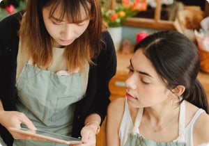 Assistentes de vendas empenhadas, elegantemente vestidas com aventais verdes, trabalham em conjunto para otimizar as operações de retalho utilizando o seu software intuitivo de gestão de retalho num tablet.