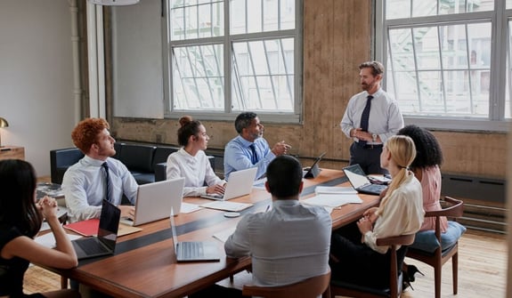 Ein Mann in Geschäftskleidung spricht sein Team an, das um einen Tisch sitzt. Effizientes Workforce Management mit Fokus auf Produktivität und Zusammenarbeit.