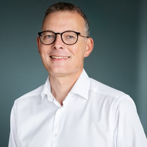 De CEO en oprichter van tamigo, Jakob Toftgaard, straalt zelfvertrouwen uit met een stralende glimlach, een scherp wit overhemd en een trendy zwarte bril tegen een betoverende blauwe achtergrond.